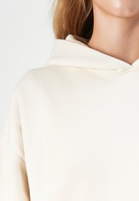 Close-up van een persoon die een crèmekleurige hoodie draagt, waarbij de halslijn, capuchon en een deel van de schouder zichtbaar zijn tegen een witte achtergrond.