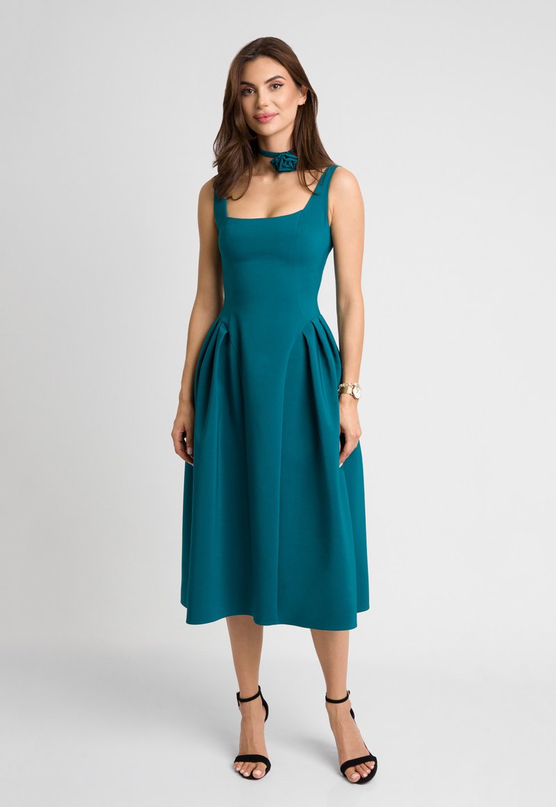 Turkusowa midi sukienka z prostokątnym dekoltem, dopasowanym górnym częścią i plisowanym dołem. Zawiera dopasowaną chokierkę z różanym detalem. Przezroczyste ramiączka i czarne obcasy.