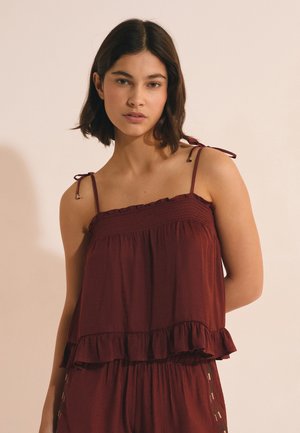 Młoda kobieta o ciemnych włosach do ramion, ubrana w bezrękawnik, marszczony bordowy top z wiązanymi na ramionach ramiączkami, stoi na jednolitym tle.