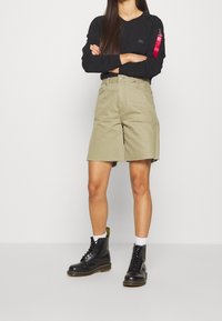 Pantalones cortos de algodón color beige con dobladillo sin terminar, combinados con una camisa de manga larga negra que cuenta con un bolsillo con cremallera. Botas negras de cordones completan el conjunto.