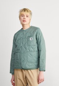 Gequilte groene jas met een ronde halslijn, drukknoopsluiting, twee voorkruiden en een logopatch op de linkerborst. Glad textuur.
