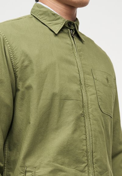 Άνδρας που φοράει χακί ζακέτα με φερμουάρ, γιακά και τσέπη στο στήθος, στέκεται μπροστά σε απλό φωτεινό φόντο.