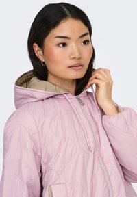 Vaaleanpunainen tikattu takki hupulla, jossa on vaaleanbeige vuori ja etutaskut. Vetoketju vetimellä ja tikatut aallokokuvioidut yksityiskohdat.