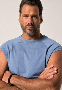 Jasnoniebieska bezrękawnik z teksturowanym wykończeniem, z okrągłym dekoltem i widocznymi detalami szwów. Model ma skrzyżowane ramiona.