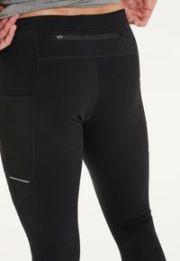 Schwarze Sportleggings mit glatter Oberfläche, ausgestattet mit einer Reißverschlusstasche auf der Rückseite und Netzpaneelen für Atmungsaktivität.