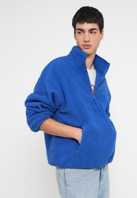 Kék polár pulóver magas gallérral, fél cipzárral az elején és oldalzsebbel. Laza szabású, texturált anyagból készült, világoskék farmerrel párosítva.