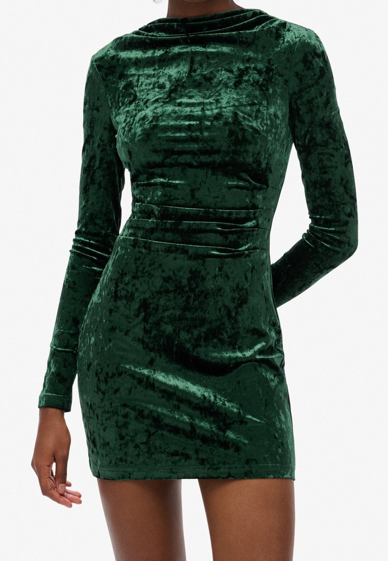 Femme portant une mini-robe ajustée à manches longues en velours froissé vert foncé, debout devant un fond uni.