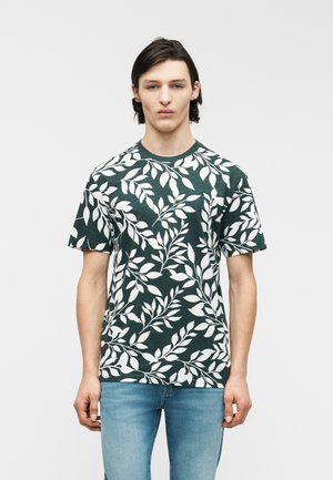 Nuori mies, joka käyttää tummanvihreää T-paitaa, jossa on valkoinen lehtikuvio, ja vaaleansiniset farkut, seisoo yksinkertaisen vaalean taustan edessä.