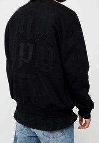 Sudadera negra de cuello redondo con texto en relieve y tonal en la parte trasera. Puños y dobladillo acanalados, ajuste relajado y textura de tejido suave.