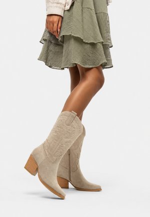 Kvinde iført lyse beige ruskindskovbojstøvler med broderi og et grønt lagdelt skørt, stående på en hvid baggrund.