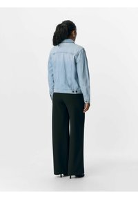 Giacca di jeans blu chiaro, con colletto classico e chiusura a bottoni, indossata sopra pantaloni neri a gamba larga. Vista da dietro, tessuto liscio.