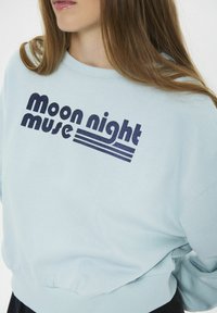 Detail zblízka na osobu, která má na sobě světle modrou mikinu se stylizovaným textem "moon night muse" vytištěným tmavě modrou barvou přes hruď.
