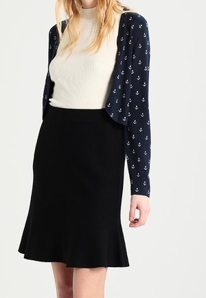 Kobieta ubrana w biały prążkowany top pod granatowym kardiganem z białymi kotwicami, w połączeniu z czarną rozkloszowaną spódnicą.