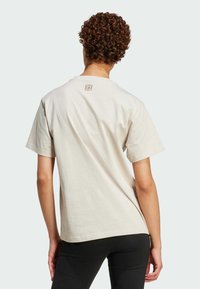 Crème-kleurig katoenen t-shirt met korte mouwen en een ronde hals, met een klein logo-detail nabij de bovenrug. Zwarte slim-fit broek zichtbaar.