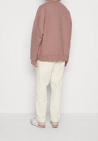 Sudadera grande de color malva con hombros caídos, combinada con pantalones crema. Zapatillas grises y rojas completan el conjunto. Textura de tela suave.