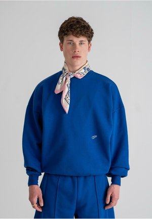 Hanorac supradimensionat de un albastru royal cu manșete cu nervuri, având un mic logo alb. Asortat cu pantaloni plisati pe albastru și o eșarfă roz cu model.