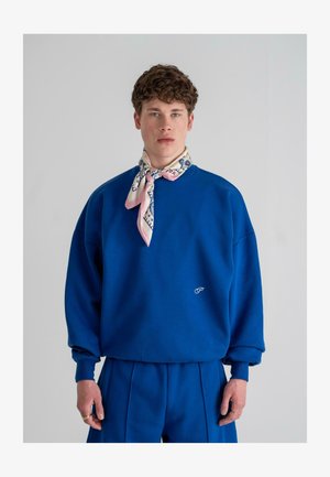 Kuninguliku sinise suuruse sweatshirt, millel on soonikuga mansetid ja väike valge logo. Kombineeritud koos vastava volditud sinise pükste ja mustrilise roosa salliga.