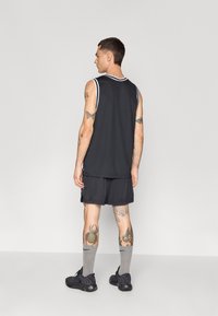 Haut de sport sans manches noir avec bord blanc, associé à un short en mesh noir. Chaussettes grises et baskets noires. Tatouages visibles sur les jambes.
