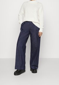 Pull en maille blanche épaisse avec motifs texturés, associé à un pantalon en denim bleu foncé ample et des chaussures à plateforme noires.