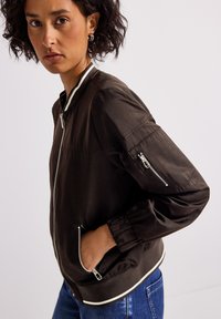 Femme portant une veste bomber noire avec des poches zippées et des bordures blanches, se tenant avec une main dans une poche et regardant droit devant.