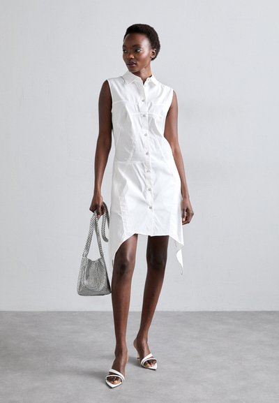 Fehér ujjatlan ingruha gombos elülső résszel, magas-lapos hem található rajta, valamint oldalsó hasítékokkal. Kiegészítve egy texturált hálós kézitáskával és pántos magassarkú cipővel.