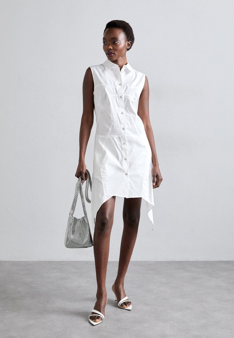 Hvid ærmeløs skjortekjole med knaplukning foran, høj-lav kant og sideslids. Accessoriseret med en struktureret mesh håndtaske og strappy hæle.