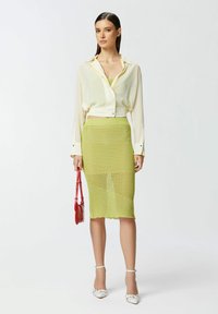 Camicia traslucida di colore crema e cut-off abbinata a una gonna verde lime a lunghezza ginocchio con texture, e borsa rossa. Sandali con tacco bianchi.