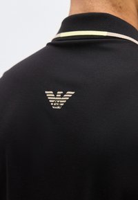 Μαύρο γιακά πόλο με λεπτές ανοιχτές ρίγες και μικρό λευκό στυλιζαρισμένο λογότυπο αετού και γράμματα "GA" στο πάνω μέρος της πλάτης.