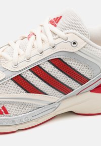 Lekki but biegowy z kremową siateczką, czerwono-czarnymi ukośnymi paskami, srebrnymi akcentami i białymi sznurowadłami. Miękka gumowa podeszwa.