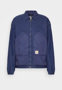 Veste en denim bleu foncé avec fermeture éclair à l'avant, grandes poches plaquées, poignets élastiques, avec une petite étiquette de marque sur la poche inférieure droite.