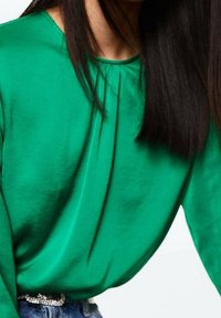 Grüne Bluse mit glatter, glänzender Textur, runder Ausschnitt und gefälteltem Designelement. Kombiniert mit einem silbernen Gürtel.