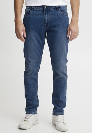 Blå denimjeans med rette ben, to frontlommer og subtil bleking. Livet har en knappelukking. Brukt med hvite joggesko.