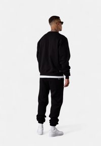 Man with short hair and neck tattoo wearing black sweatshirt, black joggers, white sneakers, and sunglasses standing facing away on white background.
