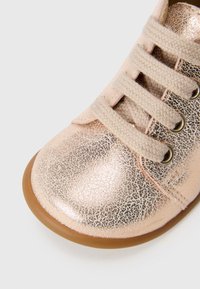 Zapato de cuero metálico rosa con una superficie texturizada, puntera redonda, cordones beige y suela de goma marrón. Presenta ojales decorativos dorados.