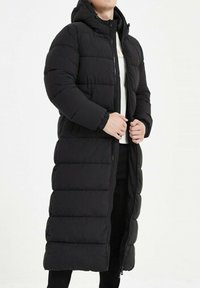 Manteau long noir rembourré avec une capuche, doté d'un design matelassé et d'une fermeture éclair. Poignets élastiques et deux poches latérales.
