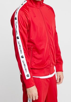 Rode trainingsjas met rits en witte Carhartt-logo strepen langs de mouw, gedragen met bijpassende rode trainingsbroek.