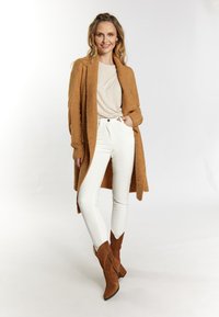 Larga chaqueta de color camel superpuesta a una blusa beige clara, combinada con jeans skinny blancos y botines de ante marrón con tacón cuadrado.