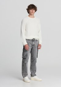 Biały sweter z prążkowanej dzianiny z długimi rękawami, zestawiony z szarymi dżinsami cargo z bocznymi kieszeniami i białymi trampkami, stojący na tle jednolitym.