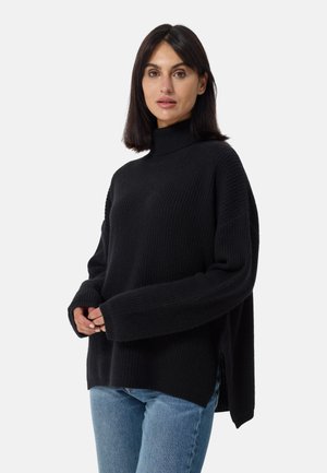 Zwarte gebreide turtleneck trui met lange mouwen, losse pasvorm, geribbelde textuur, zijsplitten, gecombineerd met lichtblauwe denim jeans.