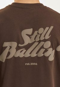 Ruskea t-paita, jossa on suuri, kohotettu vaaleanbeige teksti selässä, joka lukee "Still Ballin'" ja "est.2006" sileällä, pyöristetyllä fontilla.