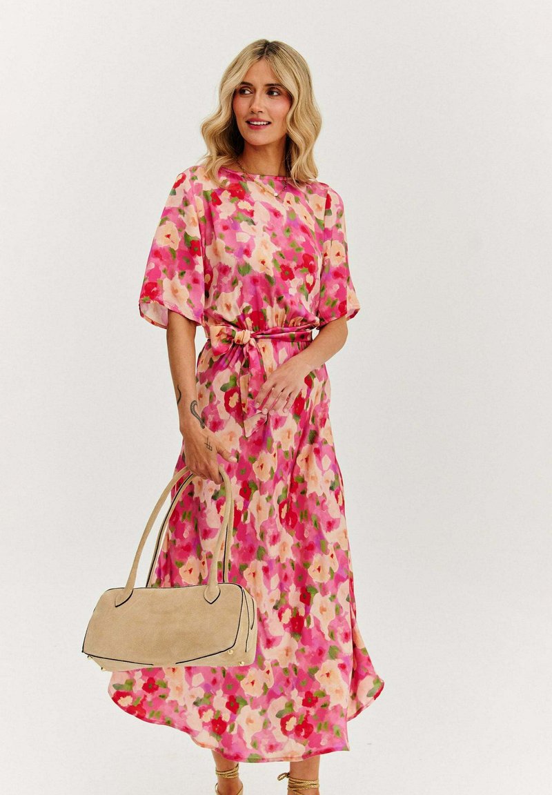 Dlouhá květinová šaty v odstínech růžové a krémové s zelenými akcenty; mají široké rukávy a mašlí v pase. Drží béžovou kabelku.