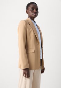 Blazer beige avec revers à cran et deux poches avant, porté avec une chemise bleu clair. Tissu lisse avec une coupe ajustée.