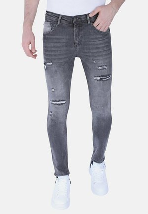Man draagt slanke, uitgebleekte grijze jeans met verweerde details en witte sneakers, poseert met één hand in de zak tegen een witte achtergrond.