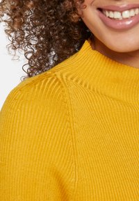 Cheveux bouclés et foncés, bouche souriante avec dents visibles, portant un pull col roulé côtelé jaune moutarde sur un fond clair.