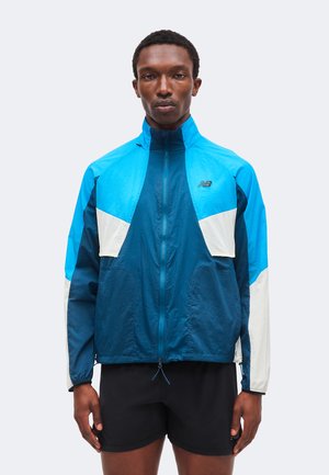 Mann trägt eine blau-türkis-weiße New Balance Zip-Jacke mit schwarzen Shorts und steht aufrecht vor einem schlichten hellen Hintergrund.