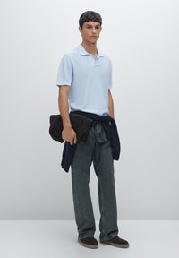 Polo shirt azzurro chiaro a maniche corte, pantaloni in denim grigi, maglione blu navy legato in vita, in mano una asciugamano marrone scuro, calzature casual.