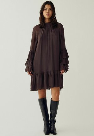 Jeune femme aux cheveux foncés portant une robe ample marron foncé avec des manches et un ourlet à volants, associée à des bottes noires montantes, debout sur un fond uni.