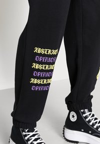 Svarta sweatpants med gult och lila tryck av "ABSTRACT OPINION" vertikalt längs med vänster ben; elastiska muddar; matchade med svarta hög-top sneakers.