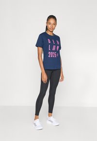 Marineblå t-shirt med fed pink tekst "BERLIN 2025", parret med sorte leggings og hvide sneakers med grå elementer. Blød bomuldsstof.