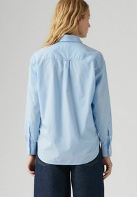 Camicetta azzurro chiaro con maniche lunghe, dettaglio della piega sul retro e orlo arrotondato. Realizzata in tessuto liscia, abbinata a denim scuro.